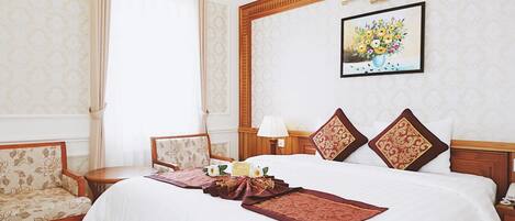 Habitación de hotel con una cama grande, dos sillas, una mesita de noche con lámpara y un cuadro con motivos florales en la pared.
