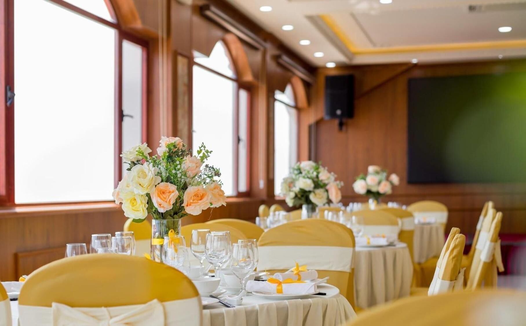 Ein Festsaal mit runden Tischen, gedeckt für eine festliche Veranstaltung, mit floralen Tischdekorationen und gelben Stuhlbändern.