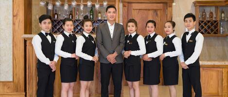 A group of hotel staff in formal attire standing in a lobby with a bar area in the background.