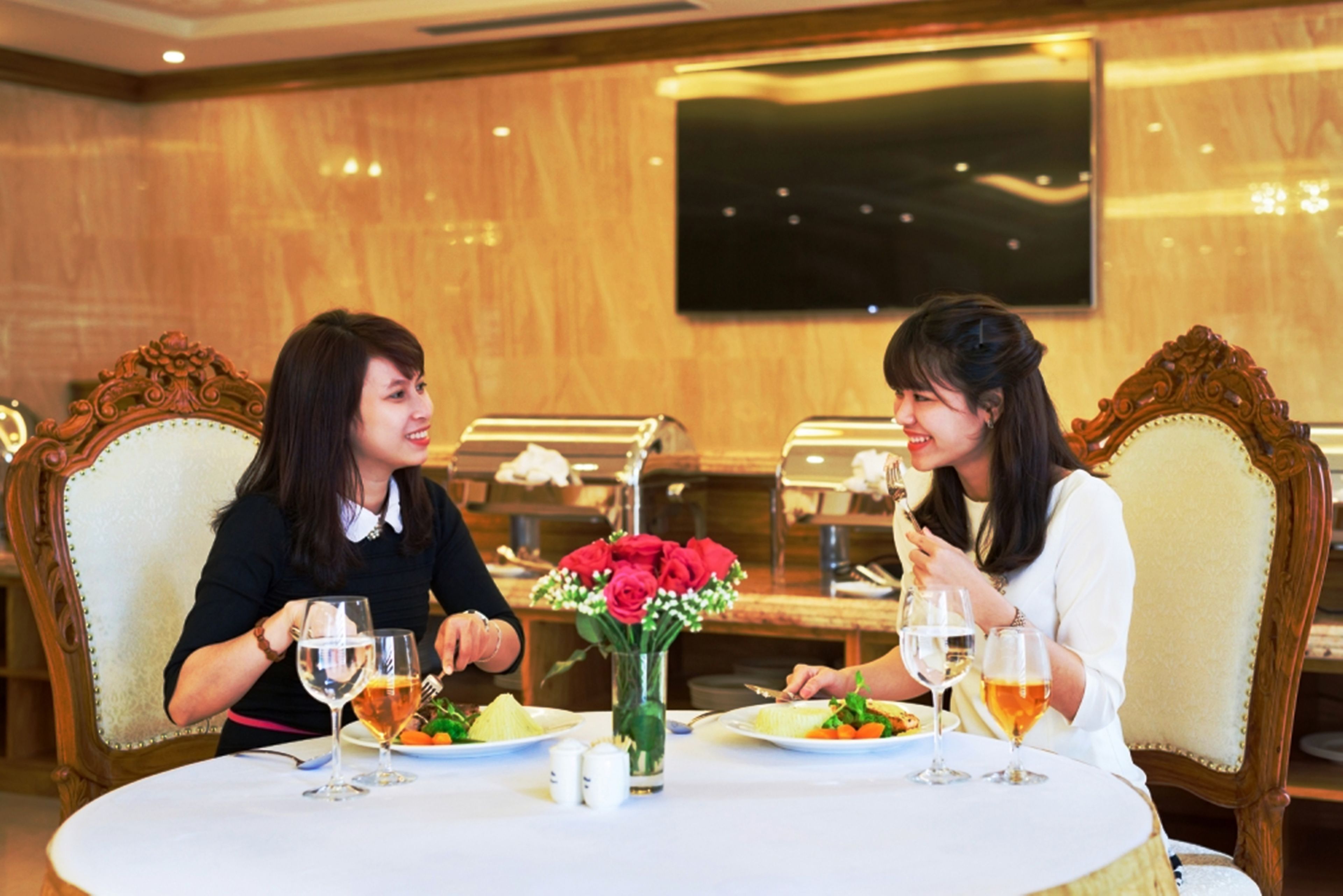 Two people dining at a table with a vase of red roses, a buffet in the background, and a television mounted on the wall.