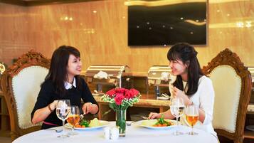 Two people dining at a table with a vase of red roses, a buffet in the background, and a television mounted on the wall.