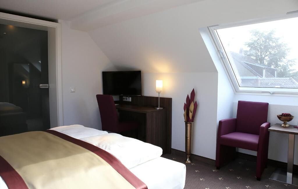 Habitación de hotel con cama, escritorio con televisor, una silla y ventana con vista a edificios.