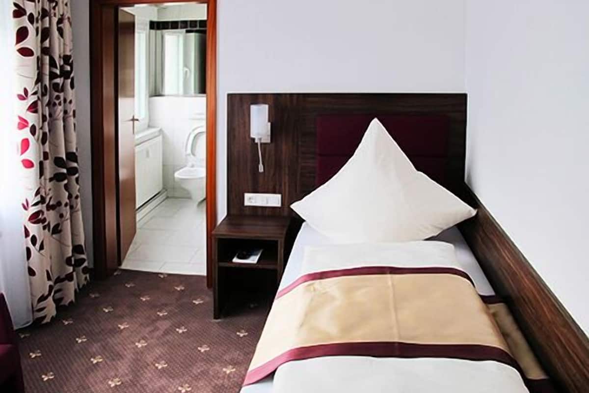 Camera d'albergo con un letto, un comodino, una testiera in legno e un bagno parzialmente visibile attraverso una porta aperta.