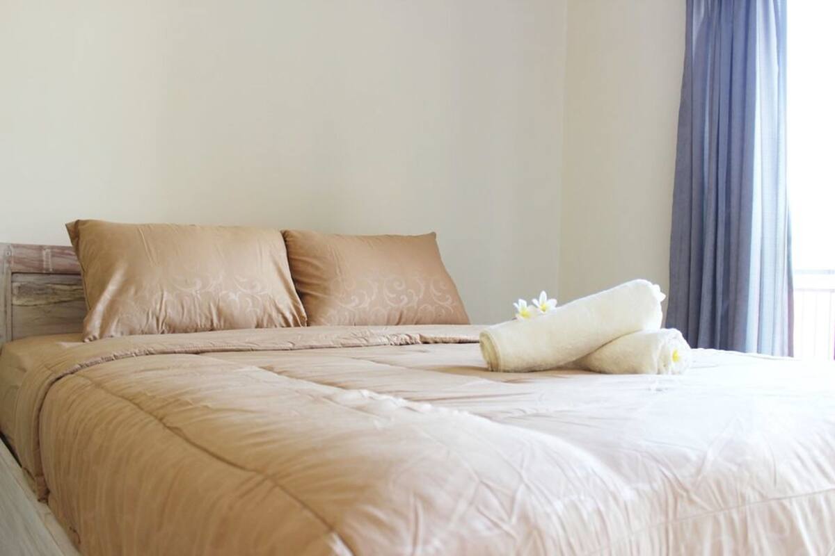 A neatly made bed with beige bedding and pillows, a rolled towel, and a flower on the bed.
