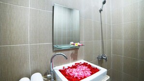A bathroom with a glass shelf, a shower, and a sink filled with rose petals. - Ratu Guest House (Kedonganan)