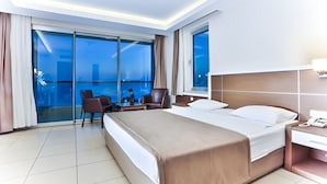 A hotel room with a large bed, a desk with a telephone, and a chair. There is a large window with curtains and a view of the city. - Venessa Beach Hotel (Alanya)