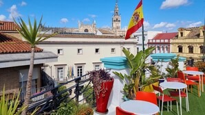 A rooftop terrace with outdoor seating, a flag, and a view of historic buildings. - Rio Hostel (Seville)