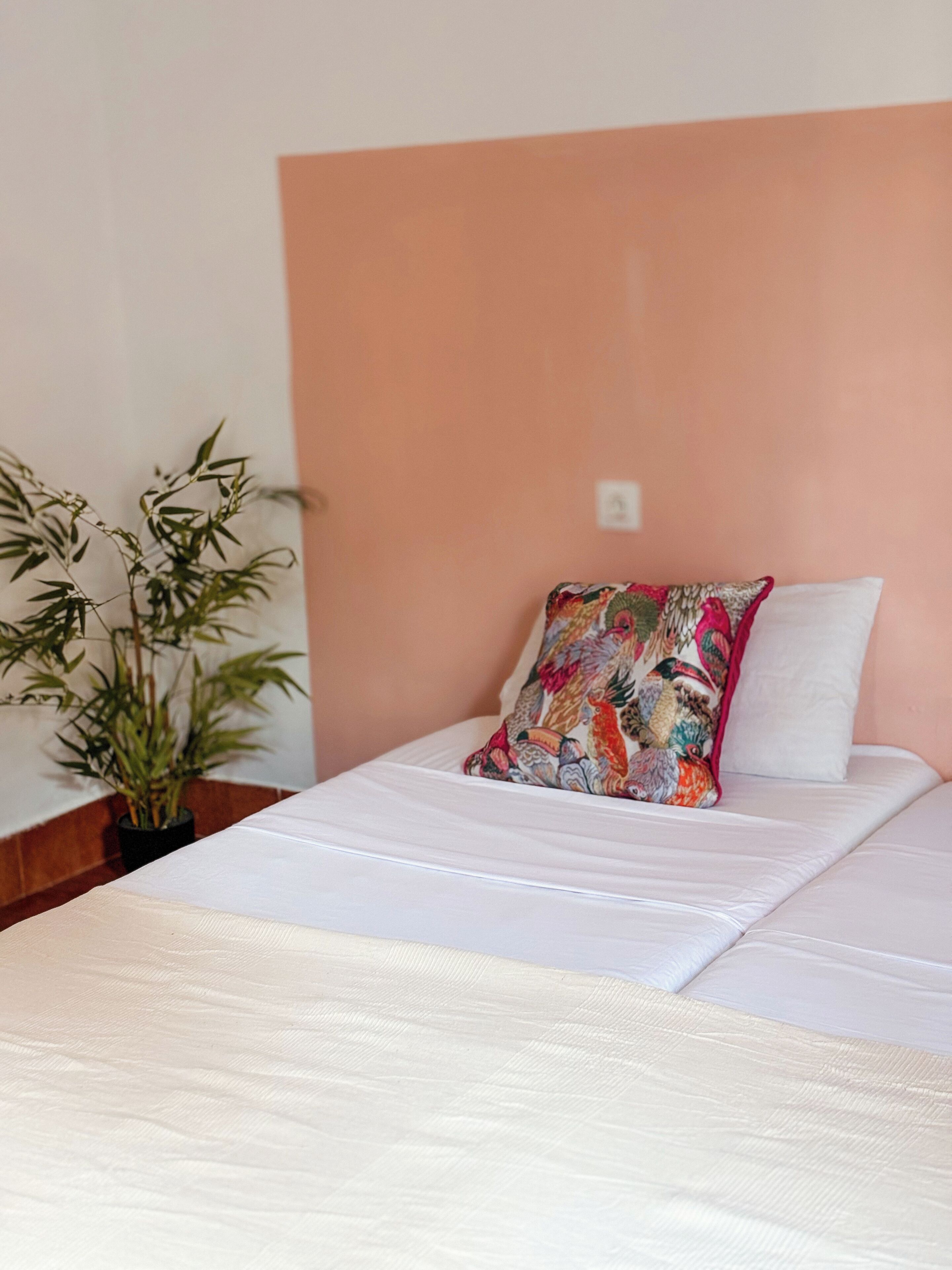 Una cama bien hecha con un colchón blanco y una almohada colorida, una planta en maceta y una pared de color melocotón.