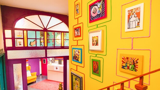 A brightly colored interior with a staircase, framed artwork on the walls, and a yellow carpet. - Rio Hostel (Seville)