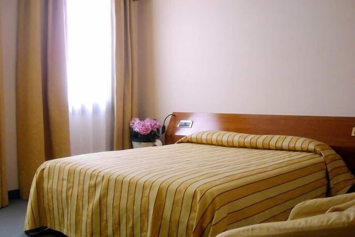 A hotel room with a bed, curtains, a nightstand, and a vase with flowers.