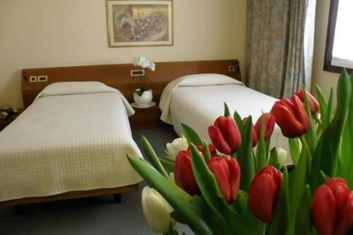 A hotel room with two beds, a nightstand with a lamp, a painting on the wall, and a window with curtains.