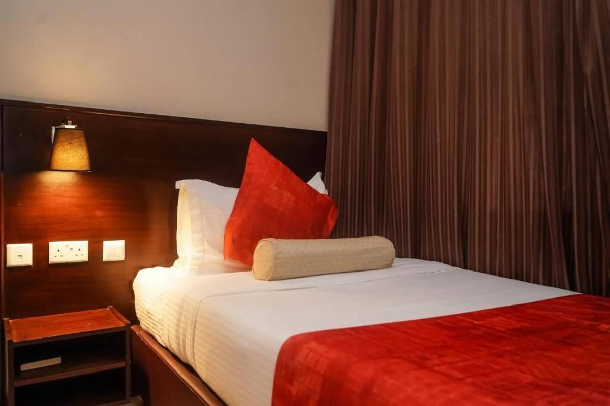A hotel room with a wooden headboard, a bedside lamp, a red throw pillow, a white pillow, a beige towel, and a red blanket.