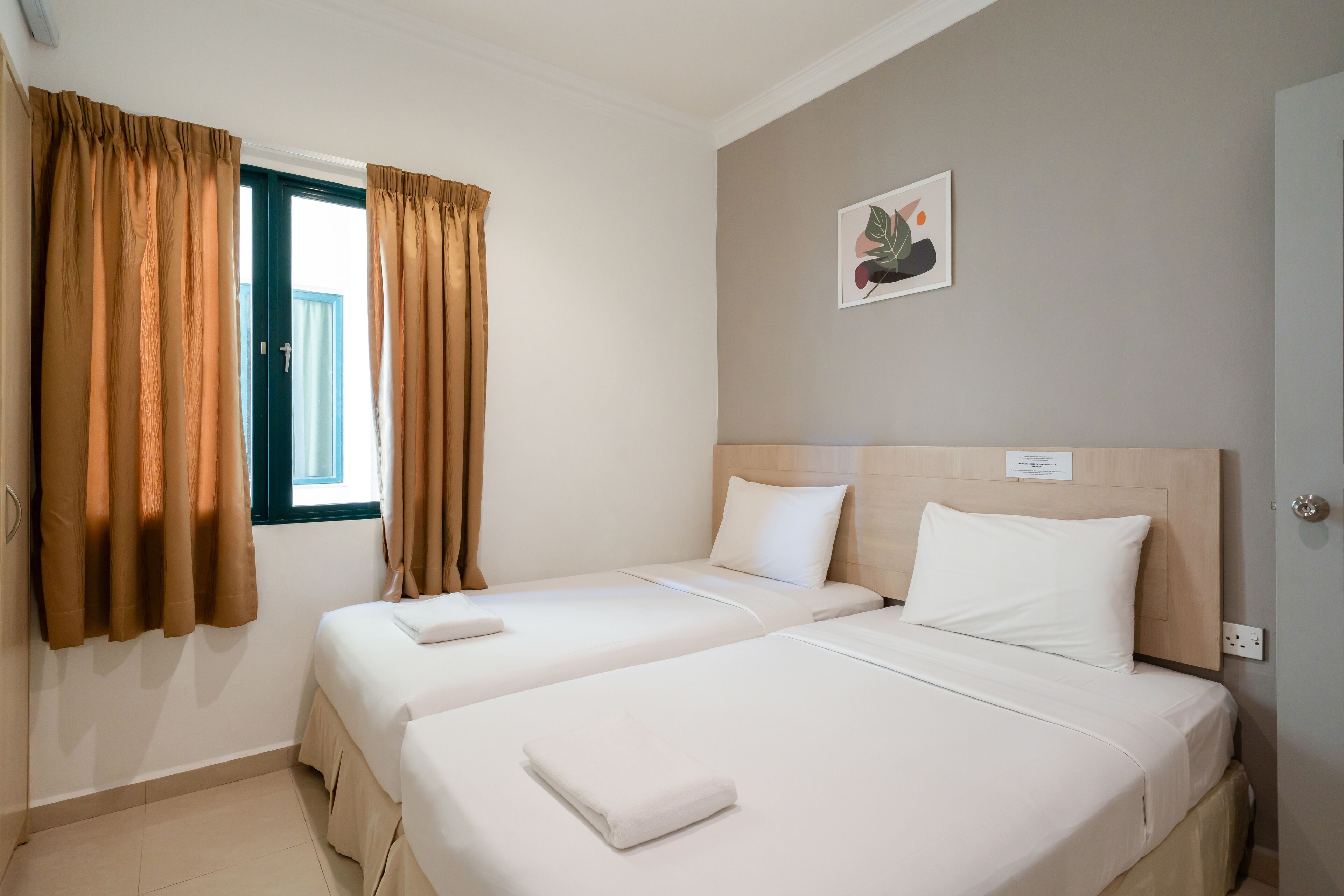 Habitación de hotel con dos camas, paredes color beige, una ventana con cortinas y una obra de arte pequeña en la pared.