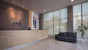 A modern reception area with a wooden reception desk, a grey sofa, and large windows offering a view of the outside. - Golden View Serviced Apartment (George Town)