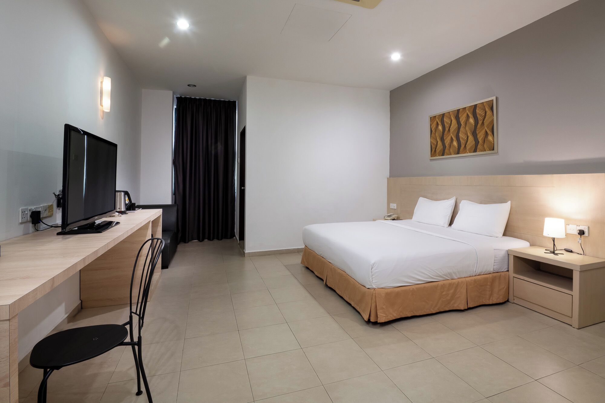 A hotel room with a bed, bedside table, lamp, desk with a computer, and a TV.