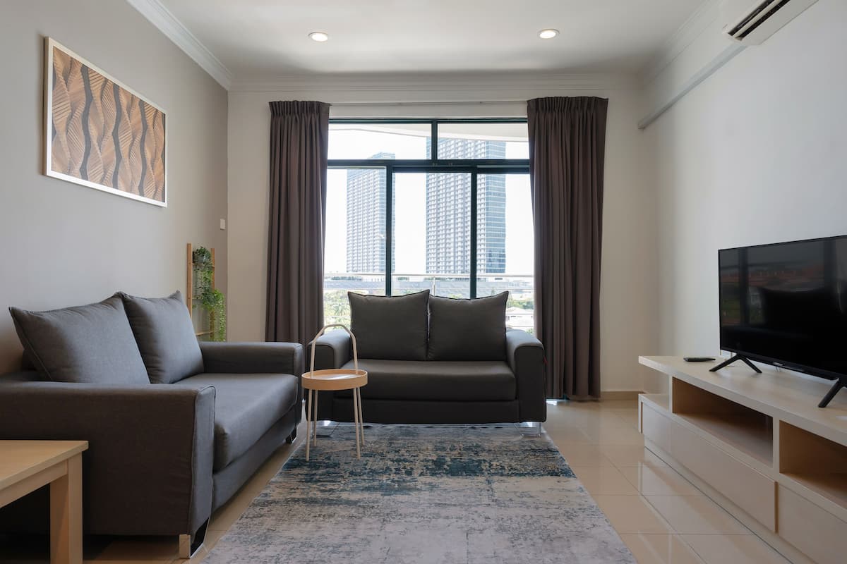 A modern living room with a grey sofa, a small side table, a flat-screen TV on a wooden stand, and a large window with curtains.