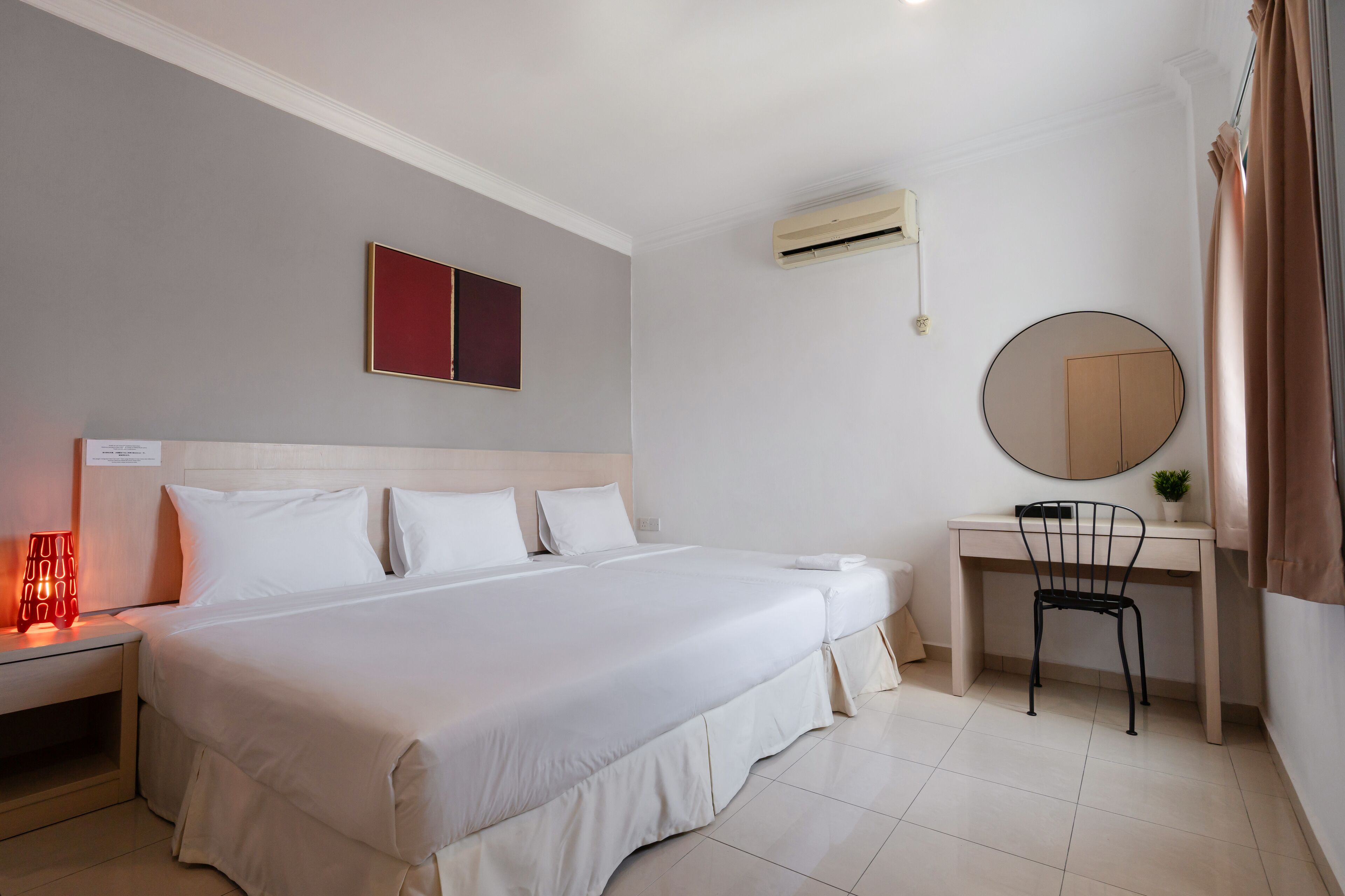 A hotel room with a large bed, a desk with a chair, a round mirror, and an air conditioning unit.
