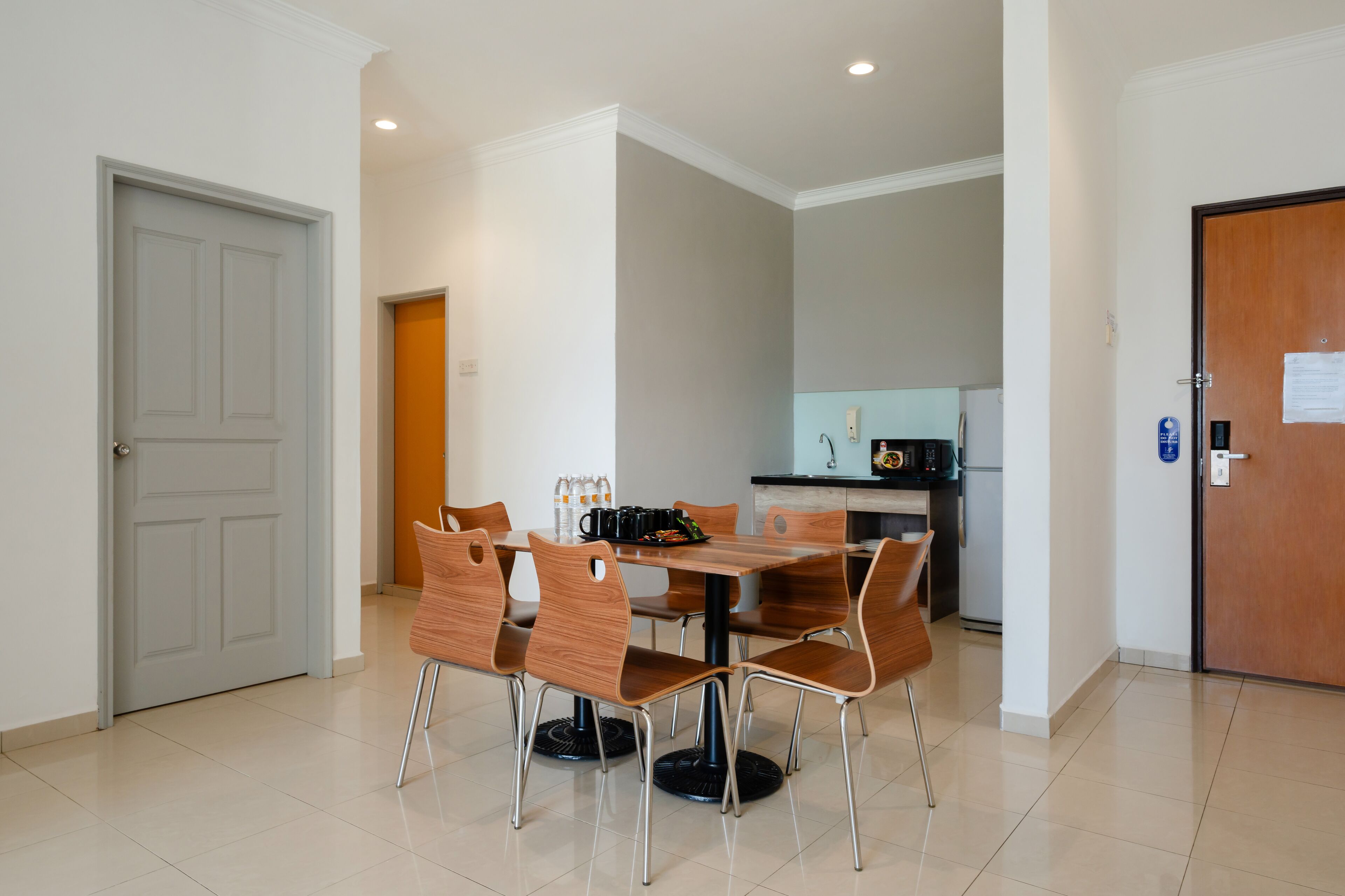 A dining area with a wooden table and chairs, a kitchen in the background, and a door on the left.