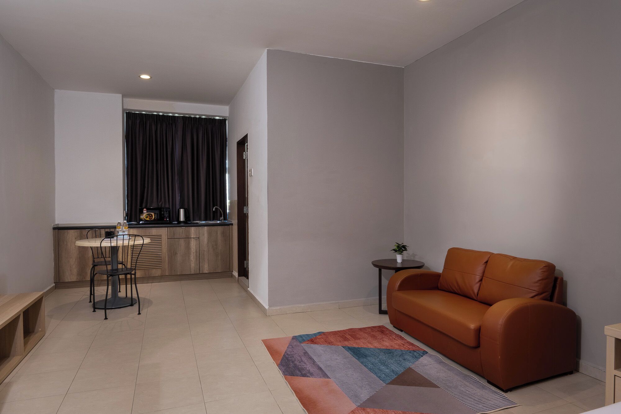 Una sala moderna con un sofá de cuero marrón, una mesita redonda y una zona de cocina con ventana y cortinas.
