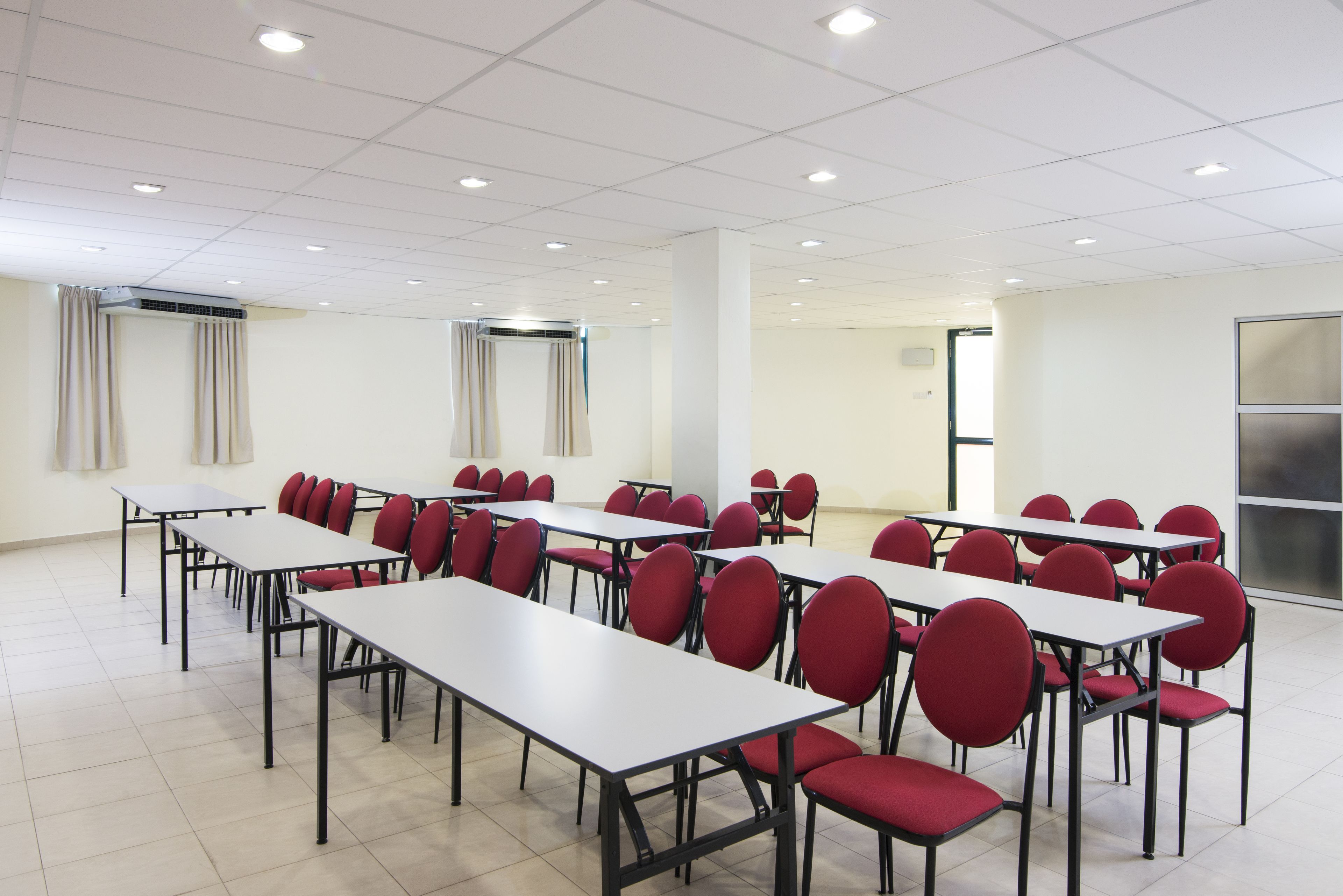 A conference room with multiple tables and red chairs, white walls, and a tiled floor.