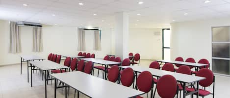 A conference room with multiple tables and red chairs, white walls, and a tiled floor.