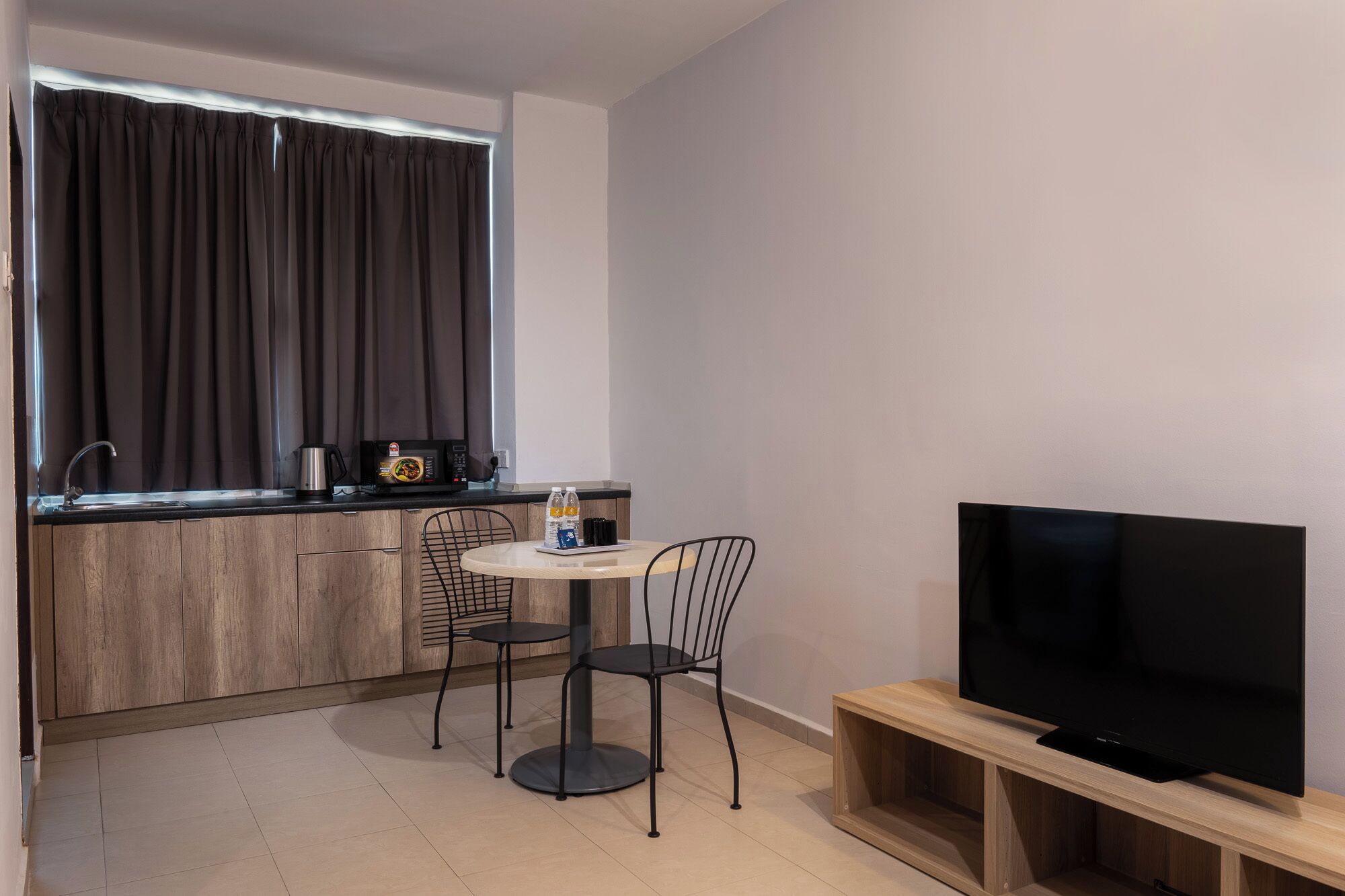 Cozinha compacta equipada com micro-ondas, chaleira e máquina de café. Há uma pequena mesa redonda com duas cadeiras, uma TV sobre um suporte de madeira e uma janela com cortinas.