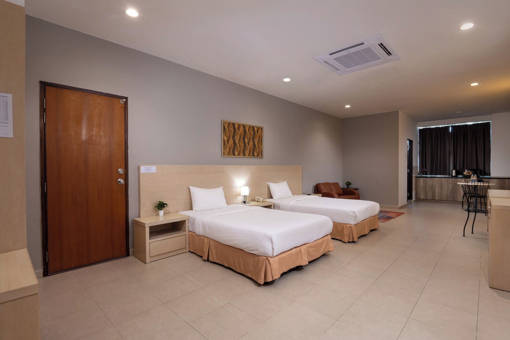 Ein Hotelzimmer mit zwei Betten, einer Holztür, einem kleinen Nachttisch mit Lampe, einer Wand-Klimaanlage und einer Küchenzeile.