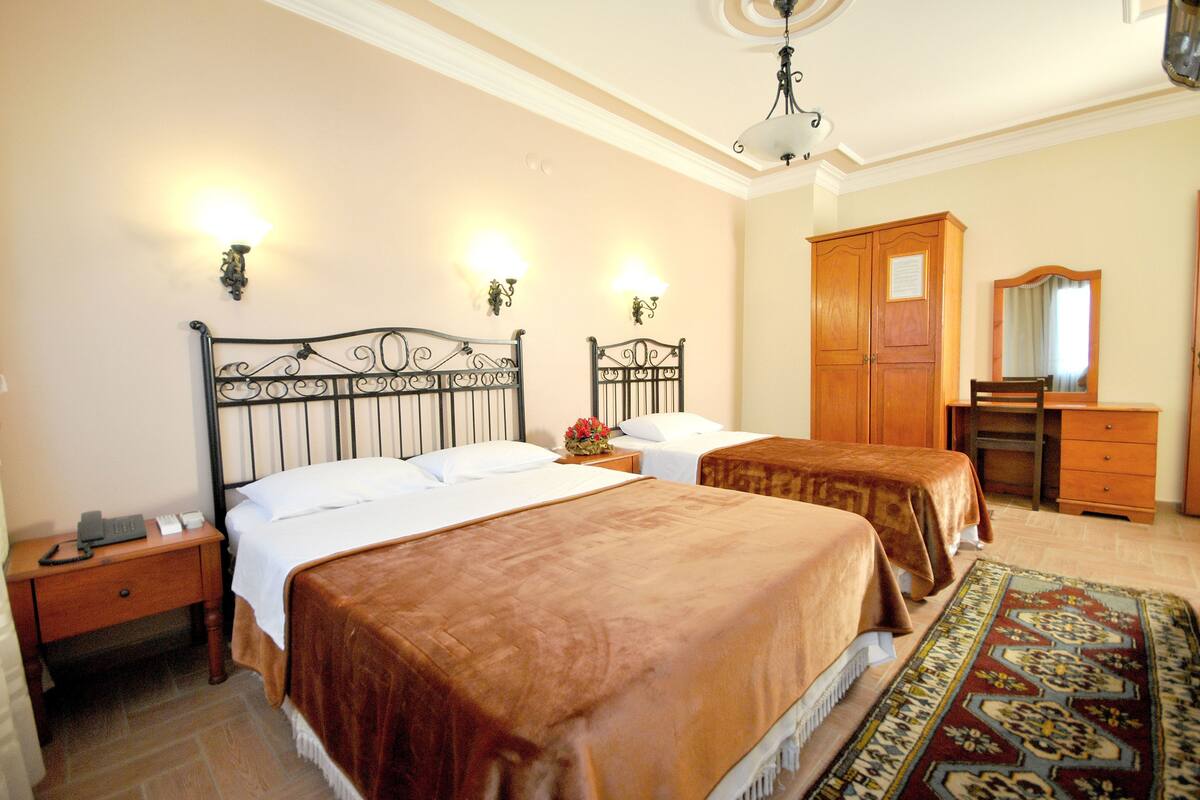 A hotel room with two beds, a wooden wardrobe, a desk, a chair, and a patterned rug.