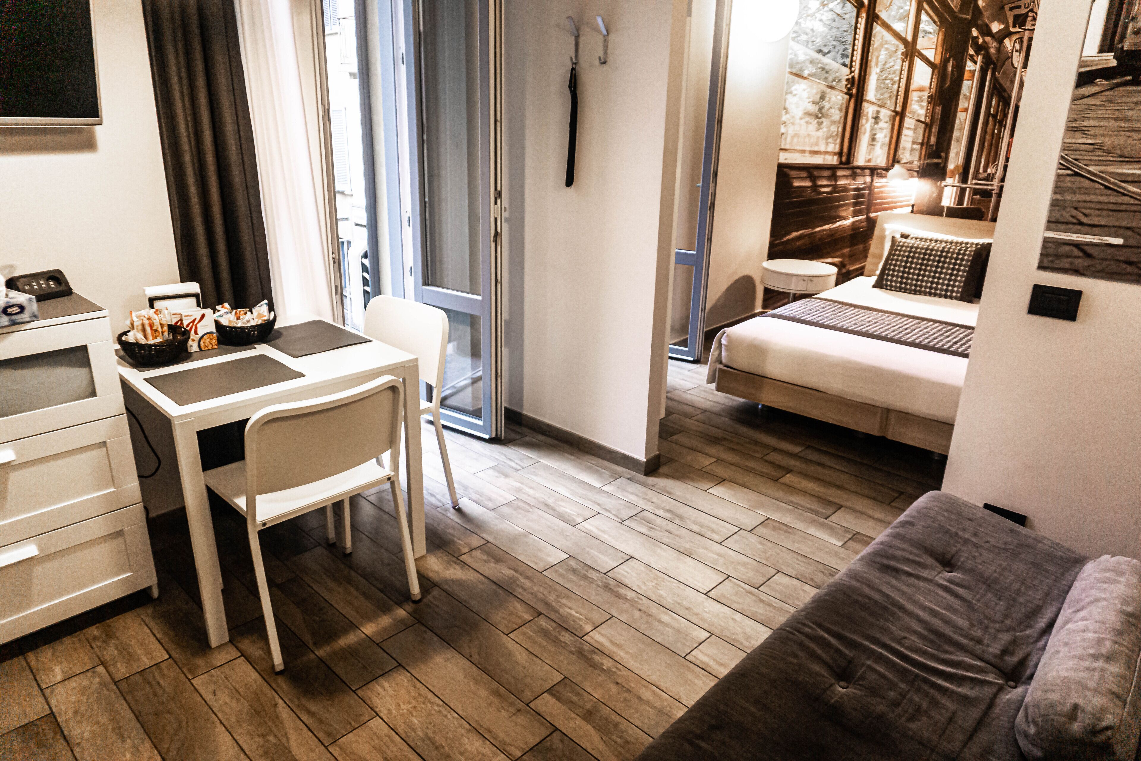 Quarto de hotel compacto com cama, pequena escrivaninha com cadeira, sofá e mesa de jantar com cadeiras.