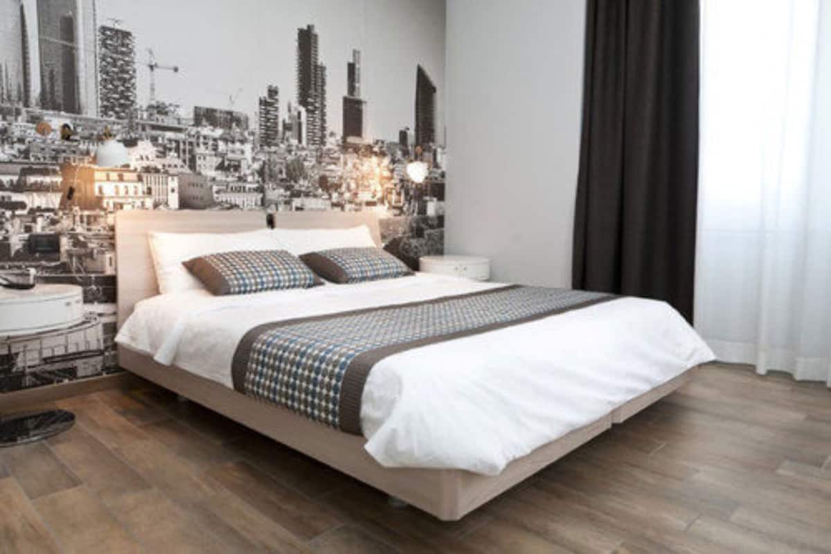 Une chambre moderne avec une fresque représentant un paysage urbain sur le mur, un grand lit recouvert d’une couvre-lit à motifs et un plancher en bois.