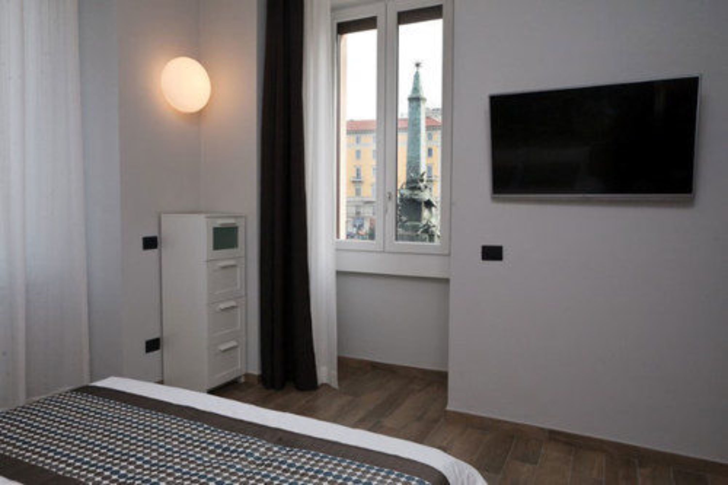 A bedroom with a bed, a wardrobe, a TV on the wall, and a window with a view of buildings.