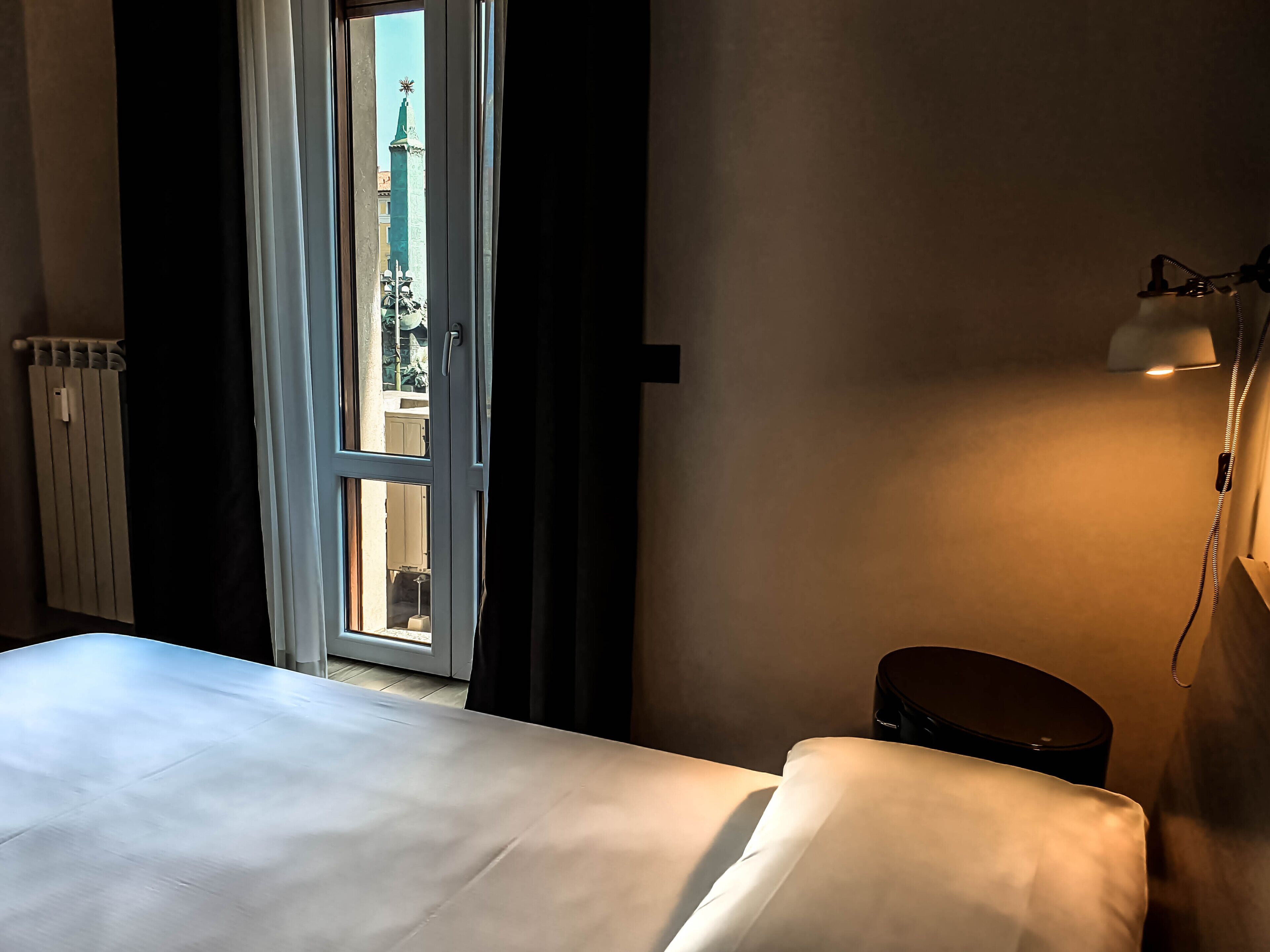 Hotelkamer met een bed, een bedlamp, een radiator en uitzicht op een gebouw door een geopende deur.