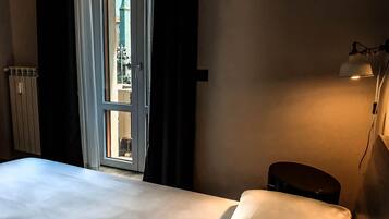 Hotelkamer met een bed, een bedlamp, een radiator en uitzicht op een gebouw door een geopende deur.