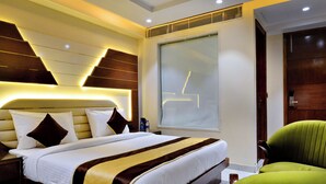 A modern hotel room with a large bed, a green armchair, and a large window with blinds.