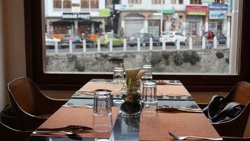 Una mesa de comedor con cubiertos y copas, un florero con una planta y vistas a una calle con coches y edificios.