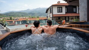 Una pareja en un jacuzzi con vistas a la montaña, barandilla de cristal y terraza de madera.