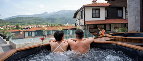 A couple in a hot tub with a mountain view, a glass balustrade, and a wooden deck.