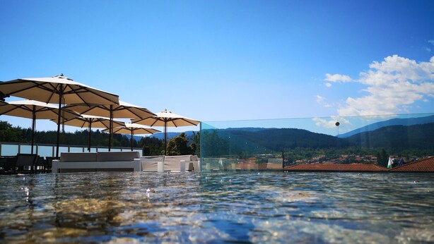 Una piscina cristalina con sillas de descanso y sombrillas, una barrera de vidrio y vistas a montañas y un pueblo al fondo.