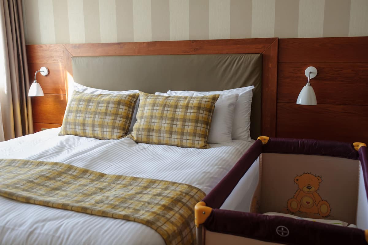 A hotel room with a large bed, two bedside lamps, and a teddy bear in a crib.