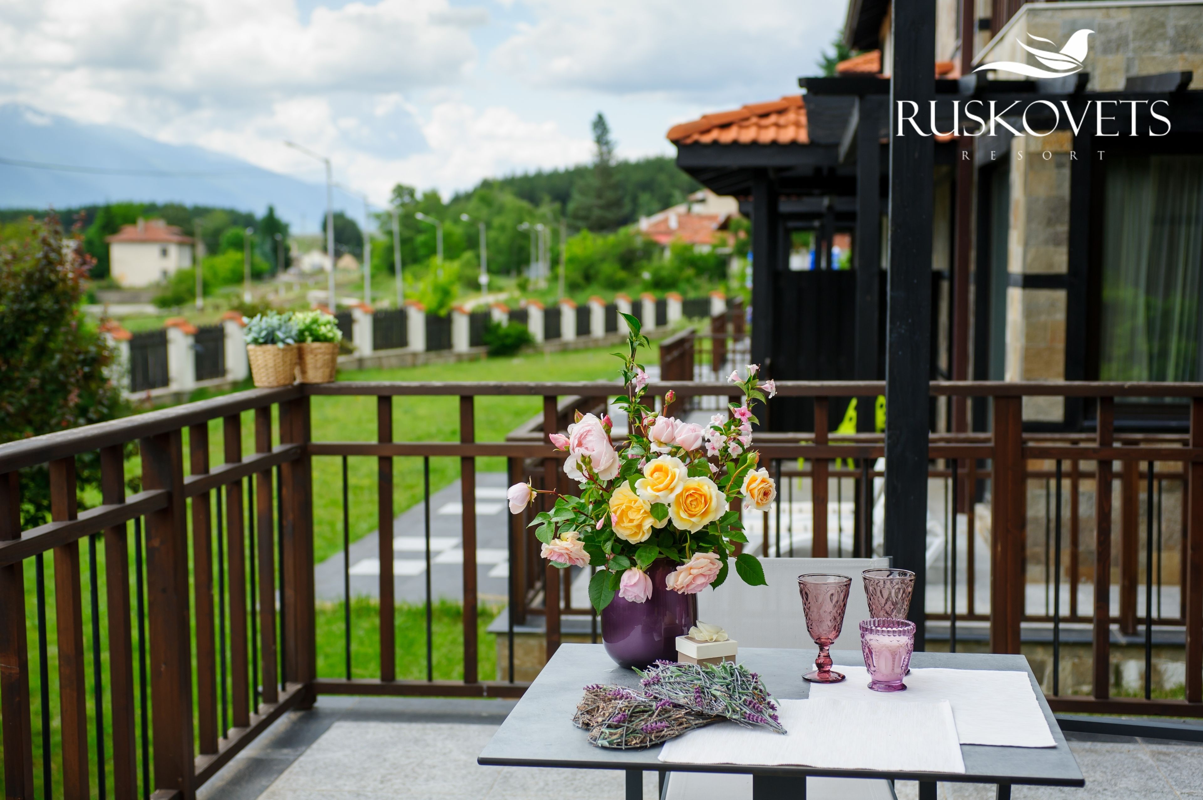 A balcony with a table set for two, a vase of flowers, and a view of a resort named RUSKOVETS.