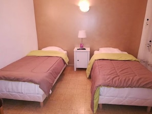 Una stanza piccola e ben illuminata con due letti singoli, ognuno con un cuscino e una coperta gialla. Tra i letti, un comodino bianco con una lampada.