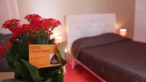 Una habitación de hotel con una cama, una mesita de noche con un arreglo floral, y un cartel que indica el nombre y la ubicación del hotel.