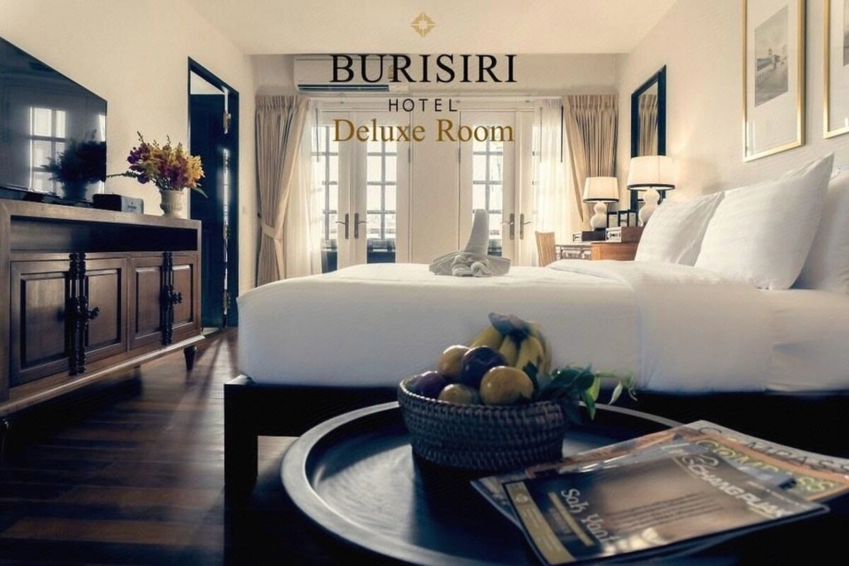 Habitación de hotel con cama, mesita de noche con frutero, una revista y televisor sobre un mueble.