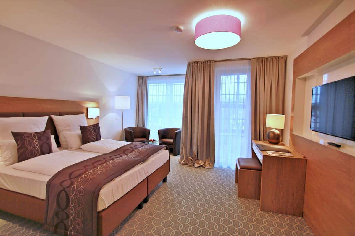 Une chambre d’hôtel avec un grand lit, deux fauteuils, un bureau avec une télévision et une fenêtre avec des rideaux.