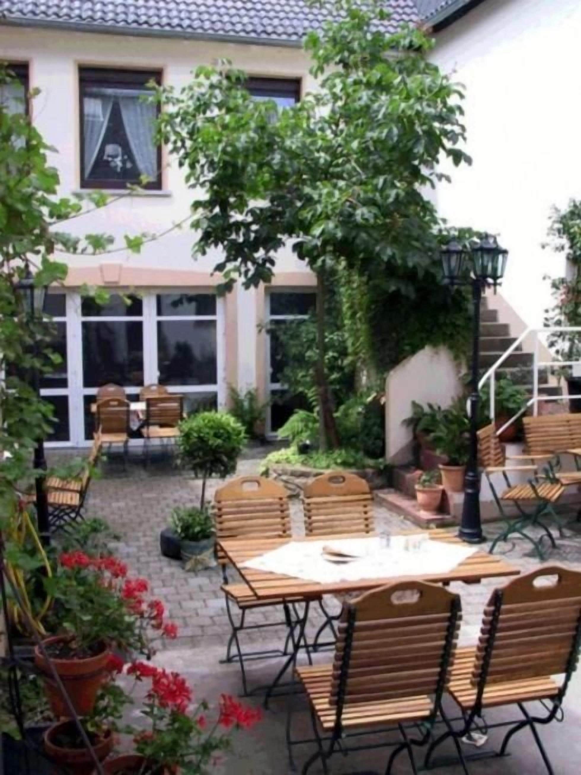 Un patio al aire libre con mesas y sillas de madera, rodeado de plantas en macetas y una escalera que conduce a un nivel superior.