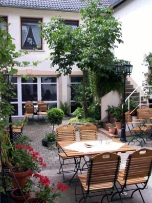 An outdoor patio with wooden tables and chairs, surrounded by potted plants and a staircase leading to an upper level.