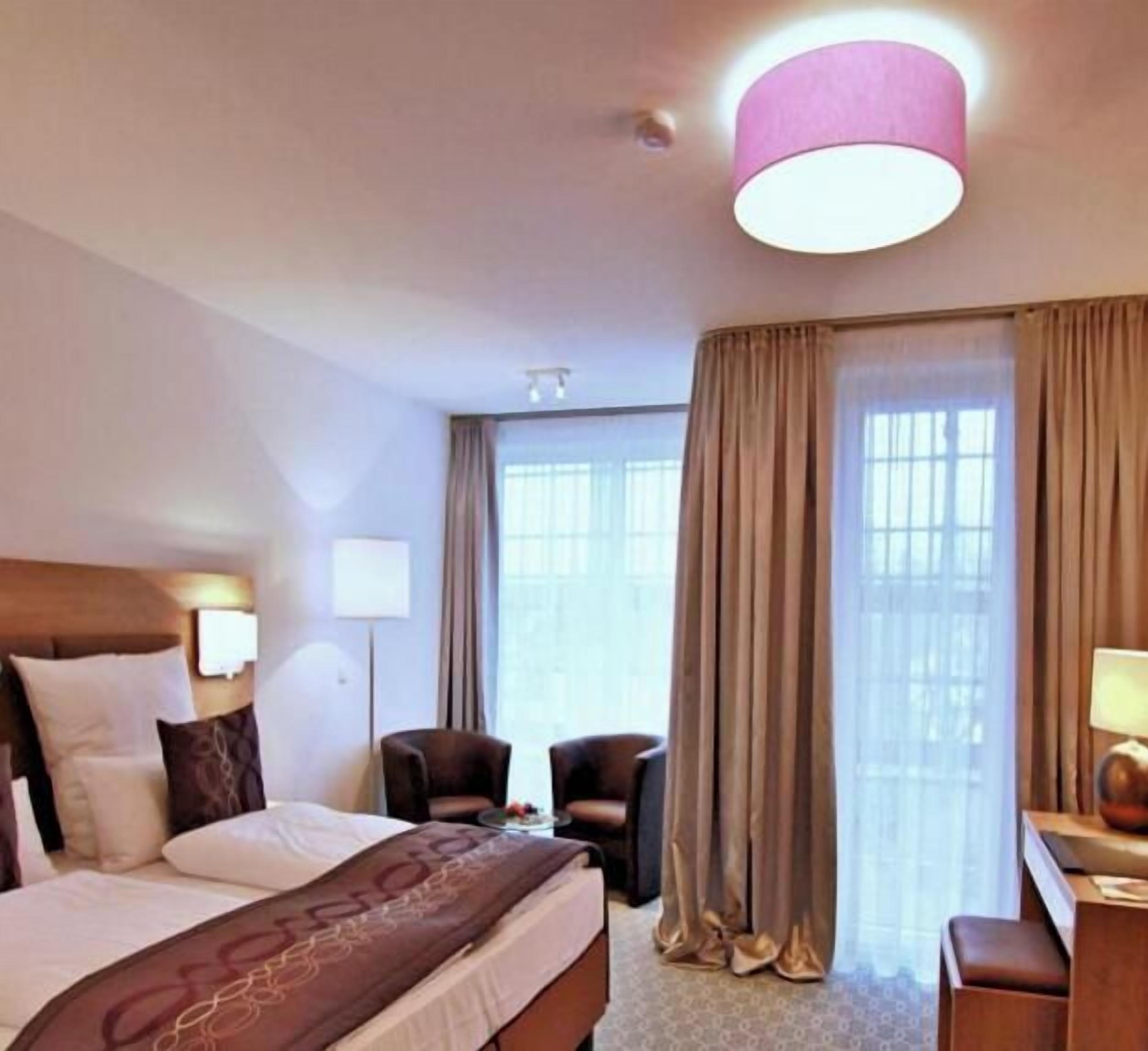 A hotel room with a bed, two chairs, a desk, and a large window with curtains.