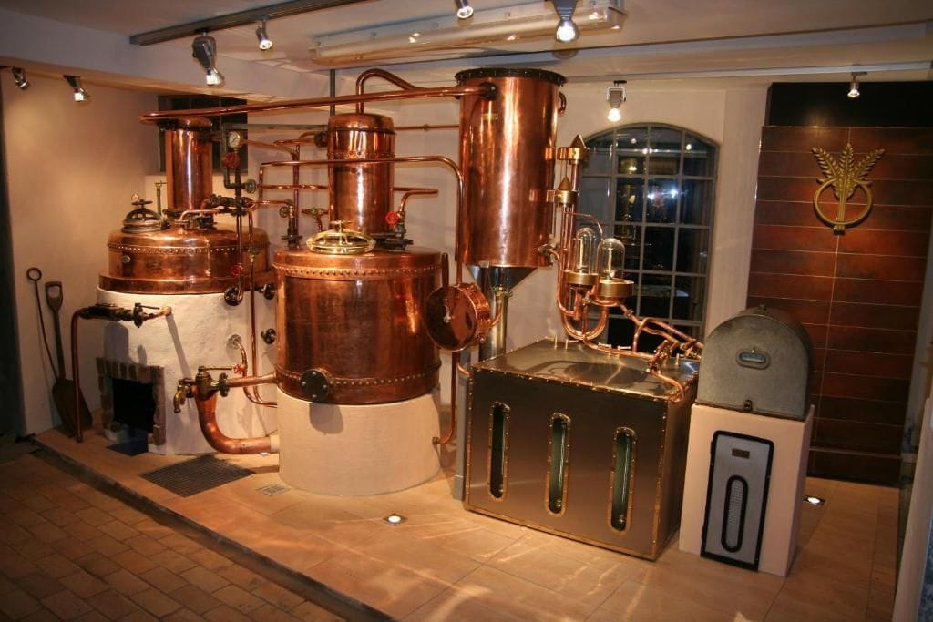 A copper distillation apparatus with glassware, a metal cabinet with ventilation slots, and a wall-mounted emblem.