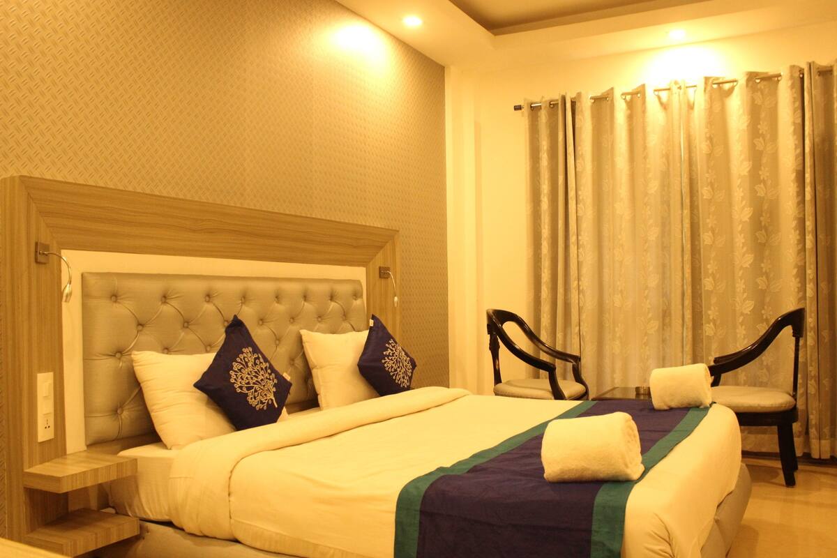 Quarto de hotel com cama grande, mesa de cabeceira, duas cadeiras e janela com cortinas.