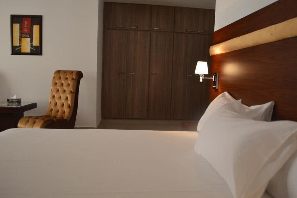 Una habitación de hotel con una cama grande, un armario de madera, un sillón capitonado y una mesita de noche con lámpara.